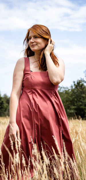 A woman with red hair wearing a dusty rose sleeveless dress stands in a field of dry grass under a blue sky with trees in the background.