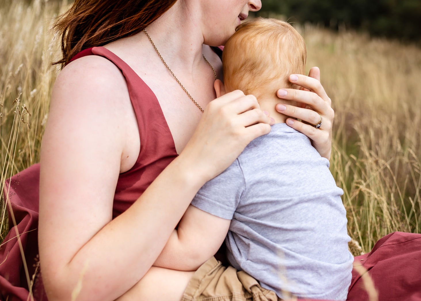 A woman with red hair sits in a grassy field holding a baby, wearing a sleeveless dusty rose top and a matching blanket.