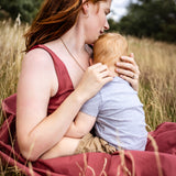 A woman with red hair sits in a grassy field holding a baby, wearing a sleeveless dusty rose top and a matching blanket.