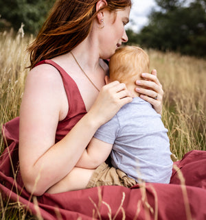 A woman with red hair sits in a grassy field holding a baby, wearing a sleeveless dusty rose top and a matching blanket.