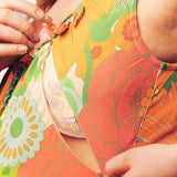 Close-up of a person adjusting a colorful floral-patterned bra or top, with hands visible and the fabric showing a mix of orange, green, yellow, and cream tones.