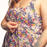 A close-up of a woman wearing a sleeveless floral dress with a purple base and multicolored flowers, adjusting the front of the dress. The background is a clean white studio backdrop.