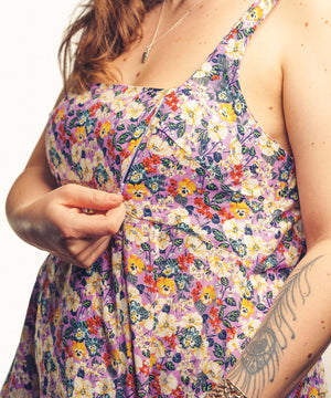 A close-up of a woman wearing a sleeveless floral dress with a purple base and multicolored flowers, adjusting the front of the dress. The background is a clean white studio backdrop.