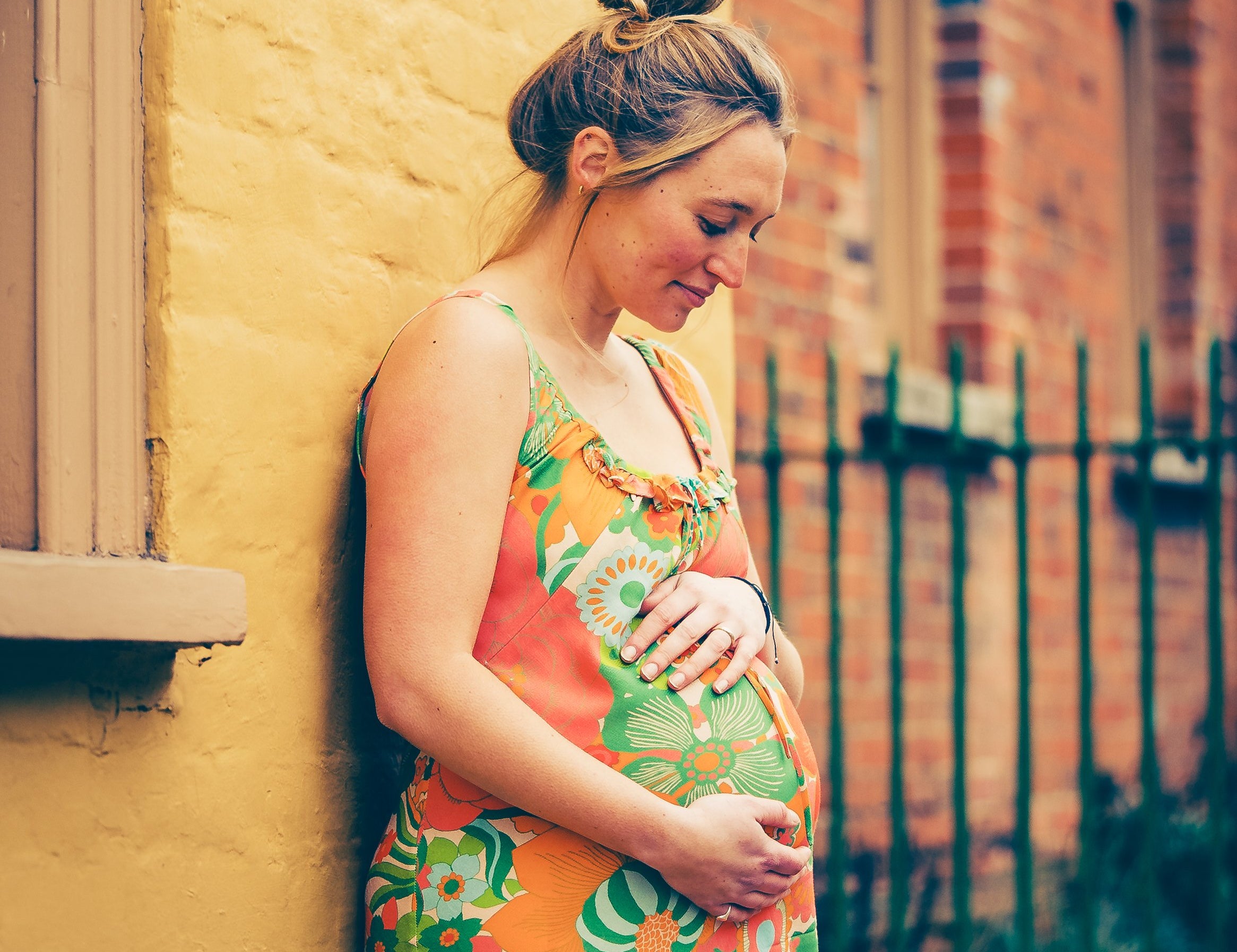Pregnant woman in a colorful dress leaning against a yellow wall.