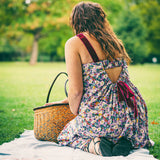 A woman sits on a white picnic blanket on a grassy park lawn, viewed from behind. She wears a sleeveless floral dress with a back cutout and dark red straps, next to a woven straw picnic basket.