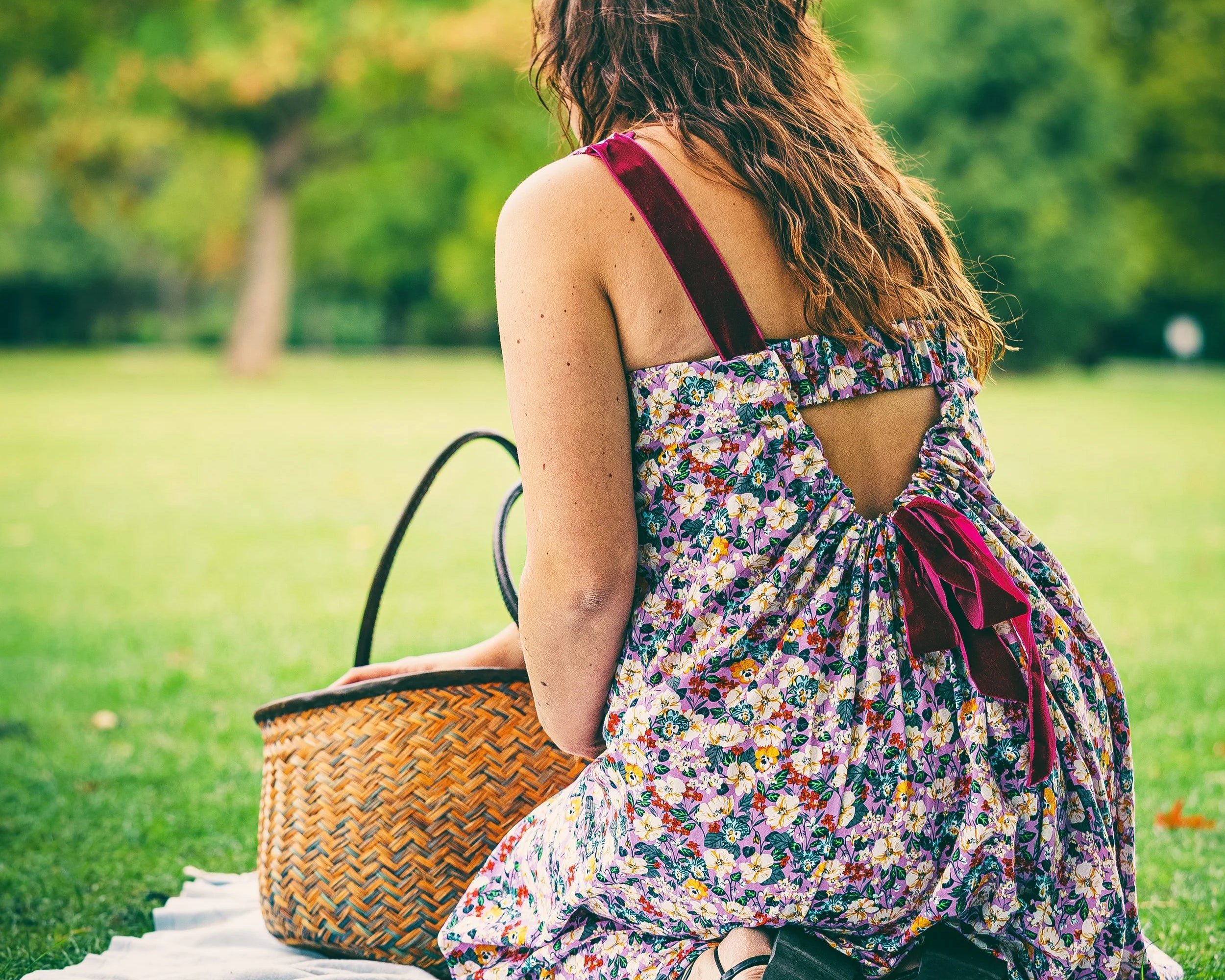 A woman sits on a white picnic blanket on a grassy park lawn, viewed from behind. She wears a sleeveless floral dress with a back cutout and dark red straps, next to a woven straw picnic basket.
