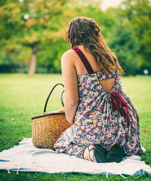 A woman sits on a white picnic blanket on a grassy park lawn, viewed from behind. She wears a sleeveless floral dress with a back cutout and dark red straps, next to a woven straw picnic basket.