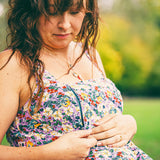 A woman outdoors wearing a floral strapless dress, looking down while adjusting the front of the dress. The background is a soft-focus green outdoor setting.
