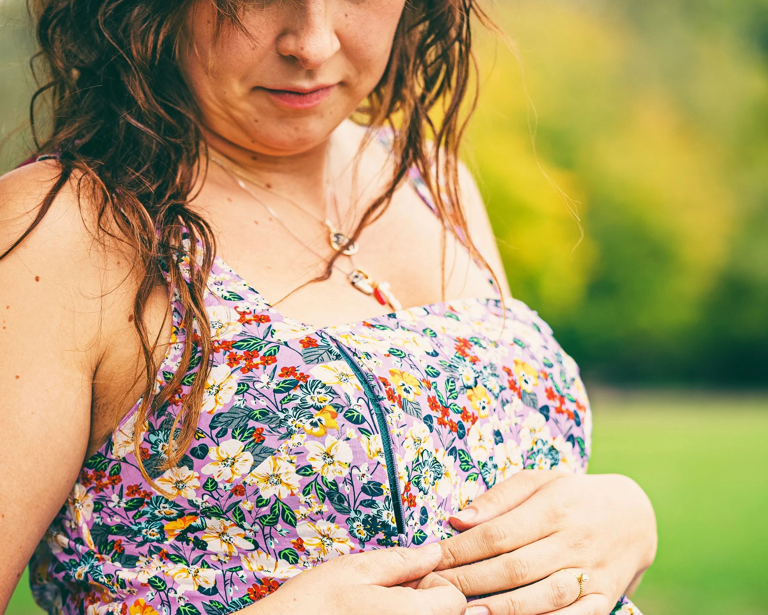 A woman outdoors wearing a floral strapless dress, looking down while adjusting the front of the dress. The background is a soft-focus green outdoor setting.