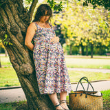A pregnant woman leans against a large tree in a park, wearing a sleeveless floral midi dress. A woven straw tote bag sits on the grass beside her.