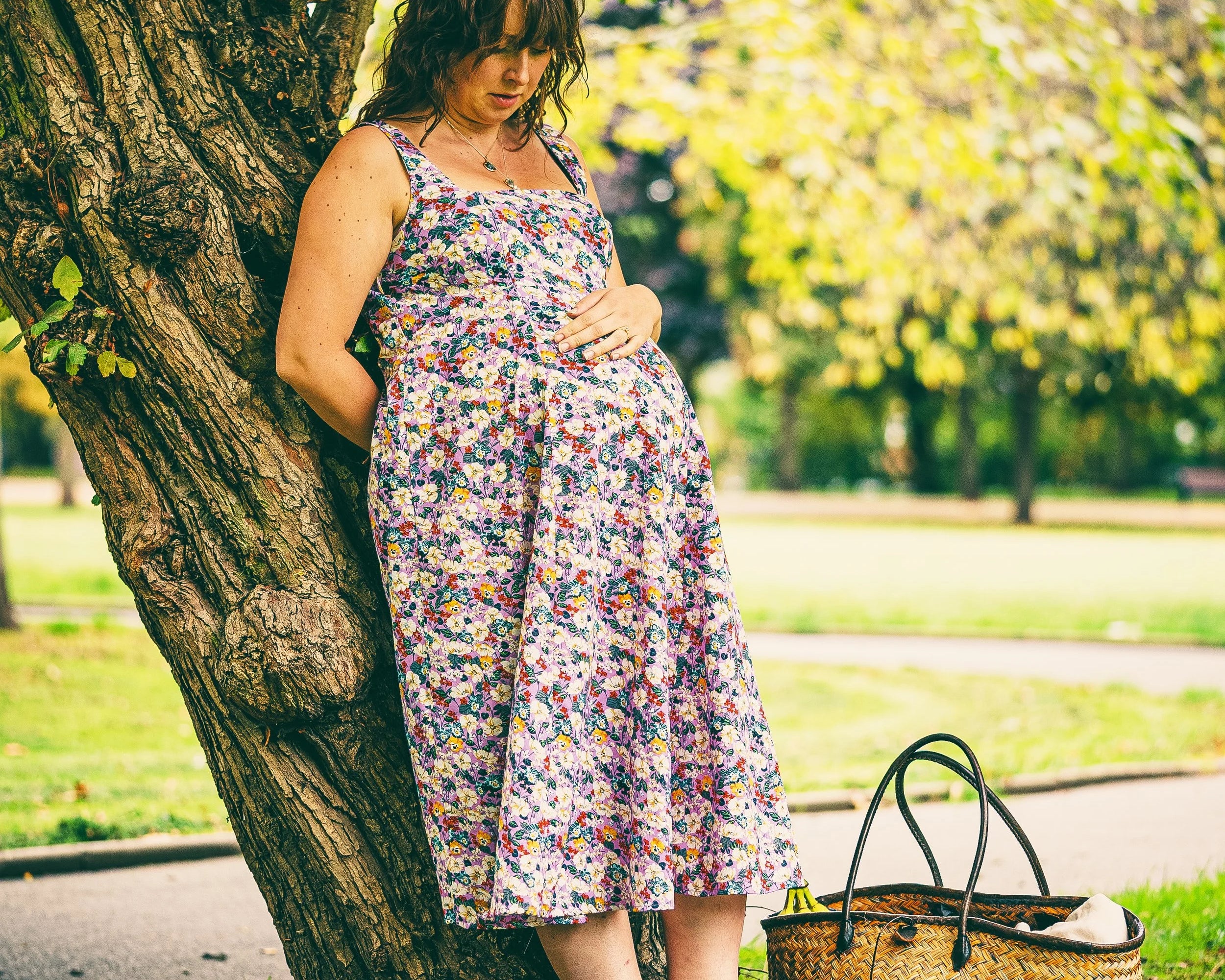 A pregnant woman leans against a large tree in a park, wearing a sleeveless floral midi dress. A woven straw tote bag sits on the grass beside her.