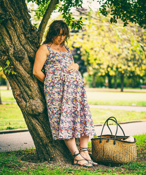A pregnant woman leans against a large tree in a park, wearing a sleeveless floral midi dress. A woven straw tote bag sits on the grass beside her.