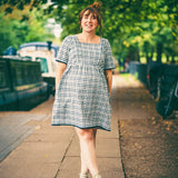 Woman in a checkered dress standing on a sidewalk with greenery in the background