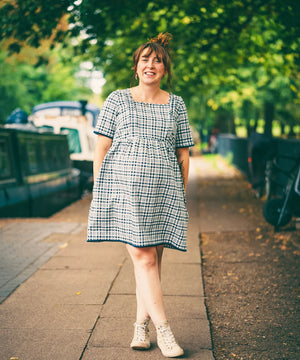 Woman in a checkered dress standing on a sidewalk with greenery in the background