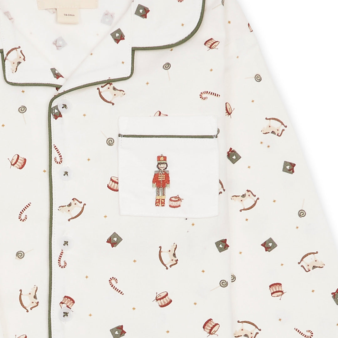 Close-up of a white long-sleeve pajama top with green piping and an all-over Christmas print featuring nutcrackers, candy canes, and small ornaments. A front chest pocket with a nutcracker embroidery is visible.