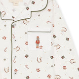 Close-up of a white long-sleeve pajama top with green piping and an all-over Christmas print featuring nutcrackers, candy canes, and small ornaments. A front chest pocket with a nutcracker embroidery is visible.