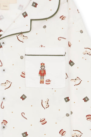 Close-up of a white long-sleeve pajama top with green piping and an all-over Christmas print featuring nutcrackers, candy canes, and small ornaments. A front chest pocket with a nutcracker embroidery is visible.