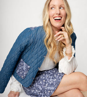 Woman wearing a blue quilted jacket and floral skirt sitting on a white chair.