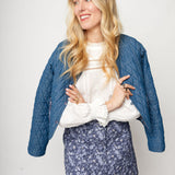Woman wearing a blue quilted jacket, white blouse, and floral skirt on a plain background