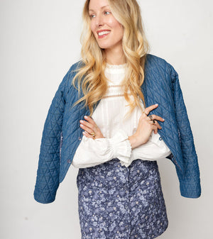 Woman wearing a blue quilted jacket, white blouse, and floral skirt on a plain background