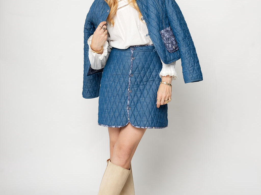Woman wearing a blue quilted jacket, white blouse, denim skirt, and beige knee-high boots on a white background