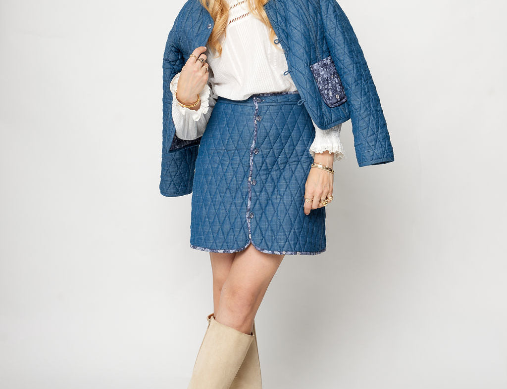 Woman wearing a blue quilted jacket, white blouse, denim skirt, and beige knee-high boots on a white background