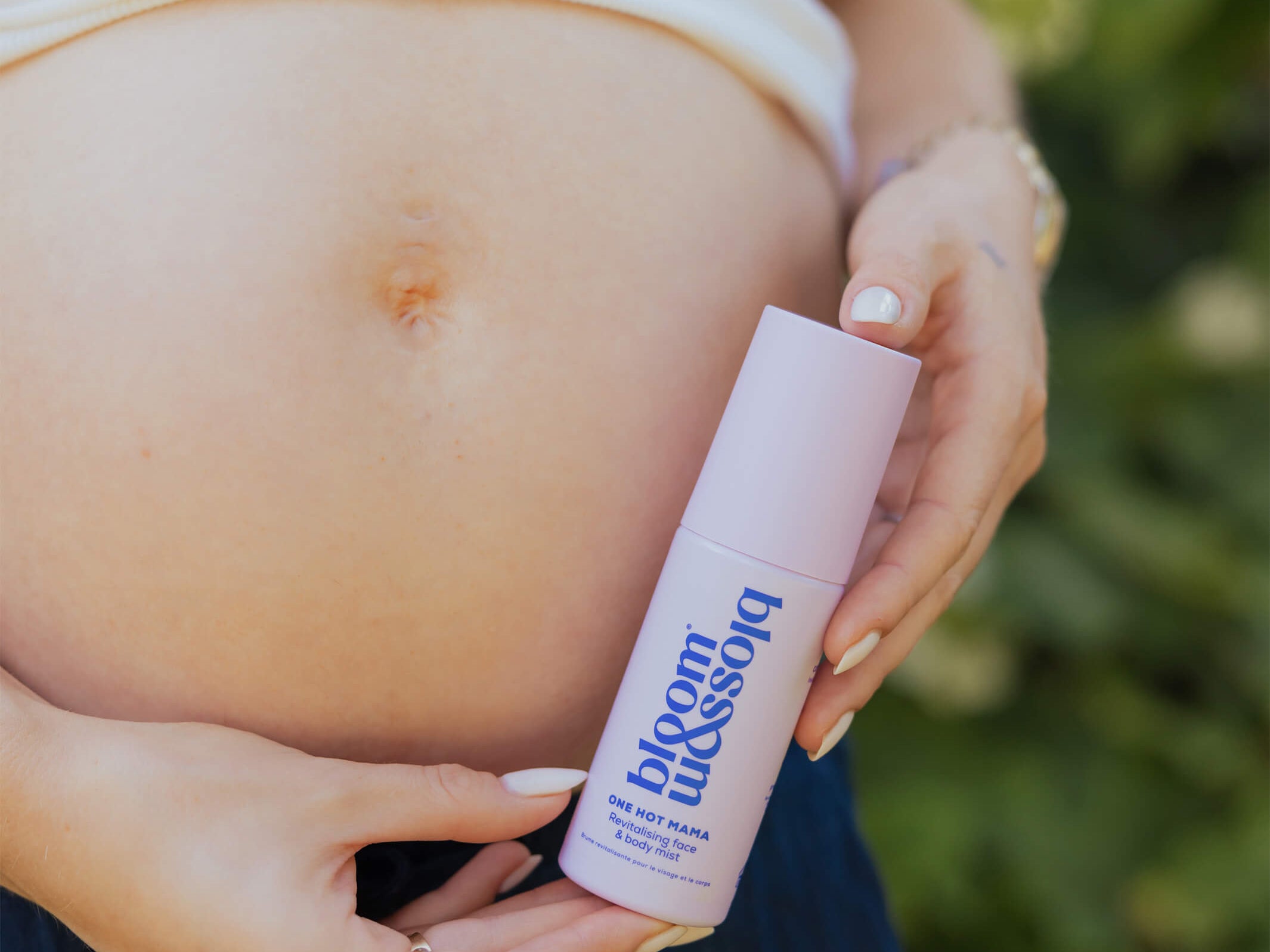 A pregnant woman holding a bottle of 'Bloom & Breathe' prenatal serum cream outdoors, with her bare stomach and hands visible against a blurred green background.