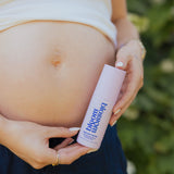 A pregnant woman holding a bottle of 'Bloom & Breathe' prenatal serum cream outdoors, with her bare stomach and hands visible against a blurred green background.