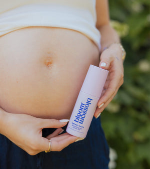 A pregnant woman holding a bottle of 'Bloom & Breathe' prenatal serum cream outdoors, with her bare stomach and hands visible against a blurred green background.