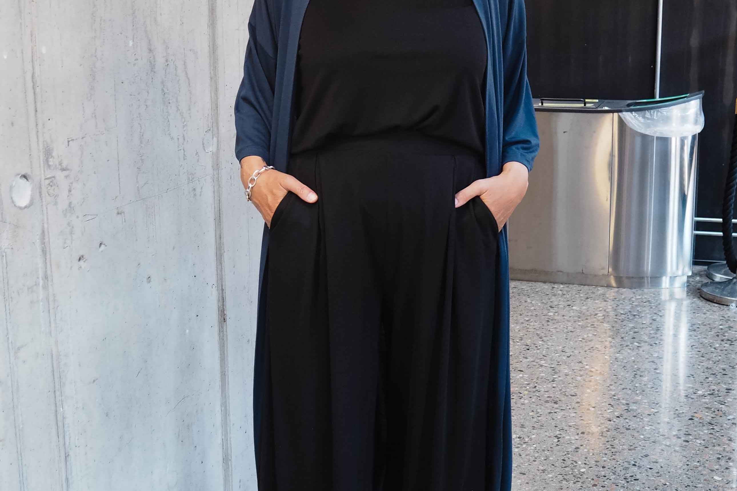 Woman in black outfit with blue cardigan standing against a concrete wall.