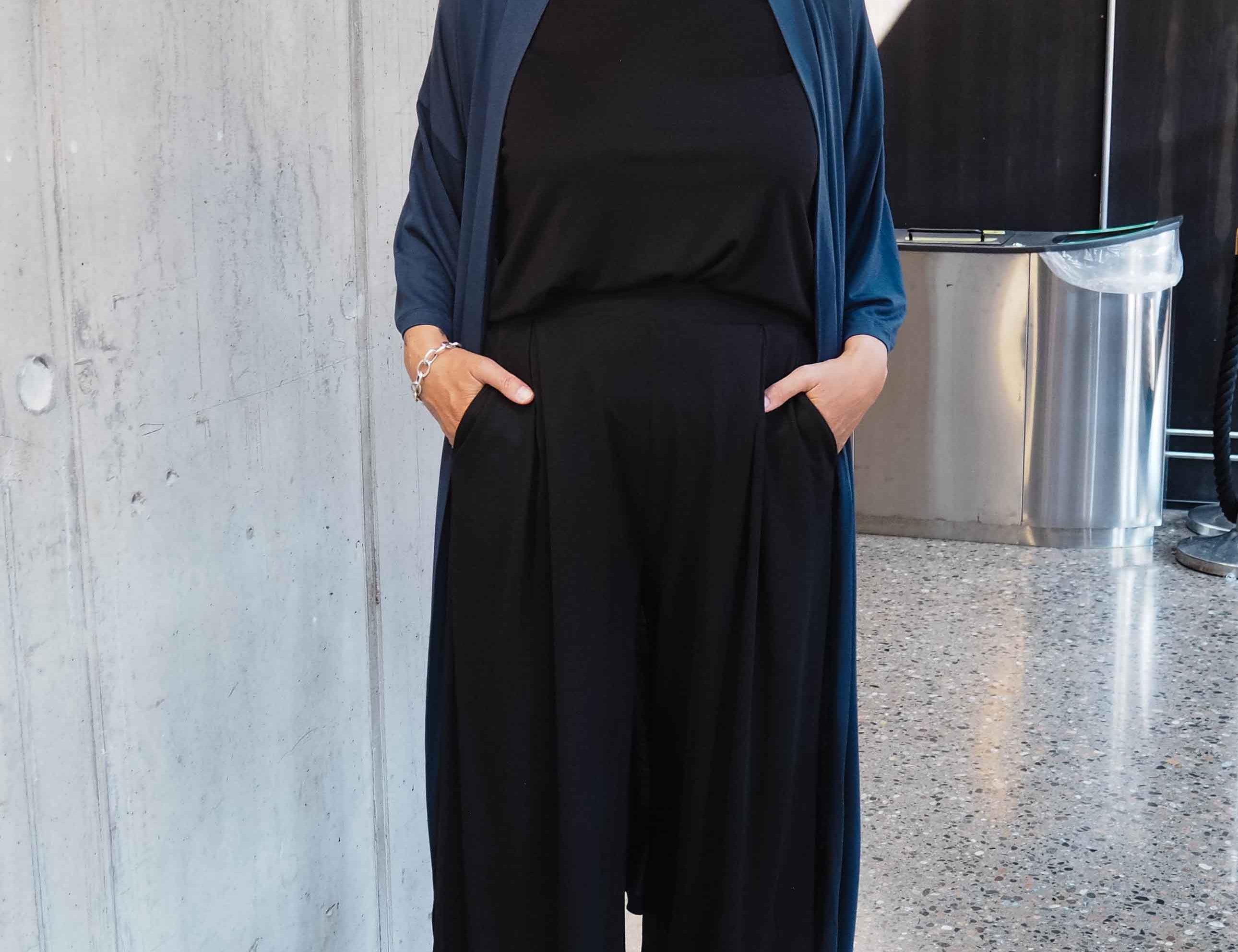 Woman in black outfit with blue cardigan standing against a concrete wall.