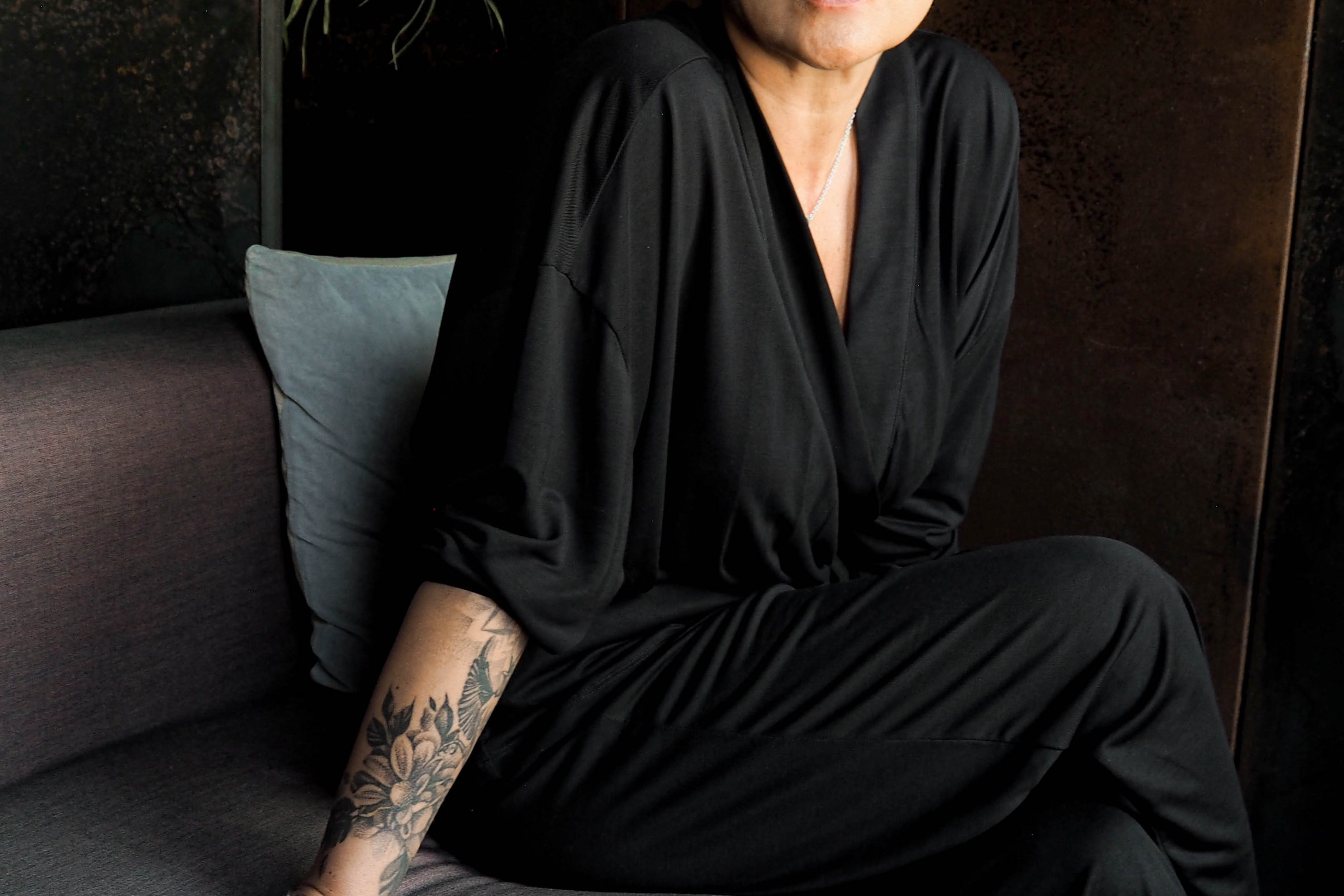 Person in a black outfit sitting on a gray couch with greenery in the background