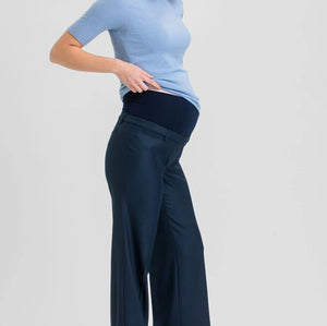 A pregnant woman posing in a light blue short-sleeve mock neck top and navy blue wide-leg maternity pants against a light gray studio background.