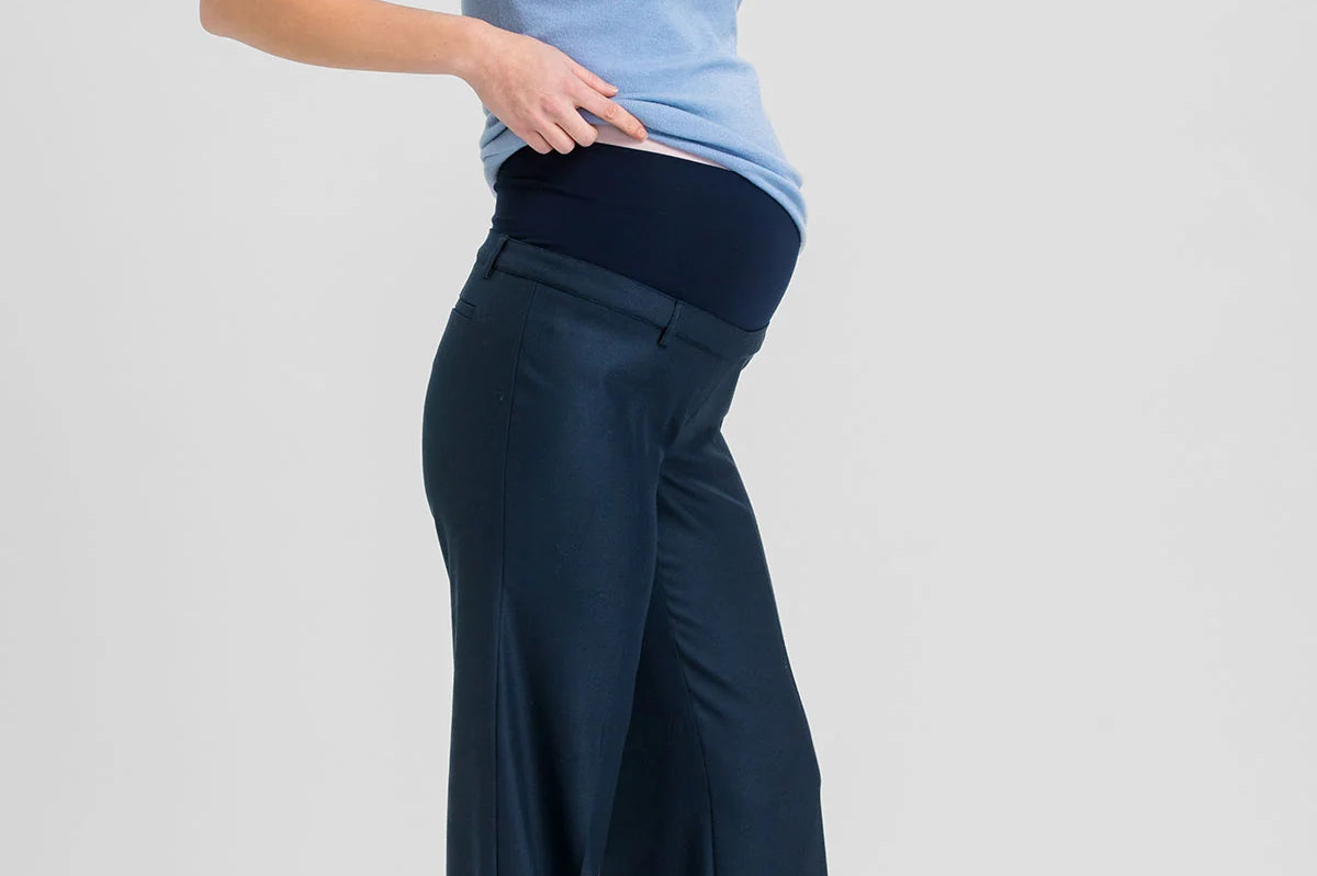 A pregnant woman posing in a light blue short-sleeve mock neck top and navy blue wide-leg maternity pants against a light gray studio background.