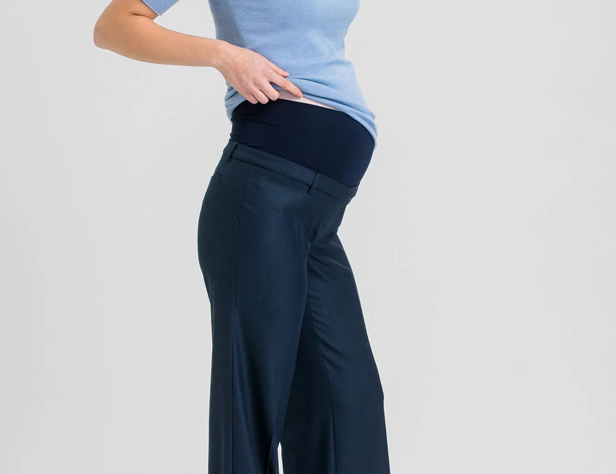 A pregnant woman posing in a light blue short-sleeve mock neck top and navy blue wide-leg maternity pants against a light gray studio background.