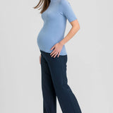 A pregnant woman posing in a light blue short-sleeve mock neck top and dark navy wide-leg trousers, paired with brown pointed-toe heels, against a solid light gray background.