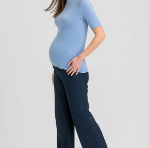 A pregnant woman posing in a light blue short-sleeve mock neck top and dark navy wide-leg trousers, paired with brown pointed-toe heels, against a solid light gray background.
