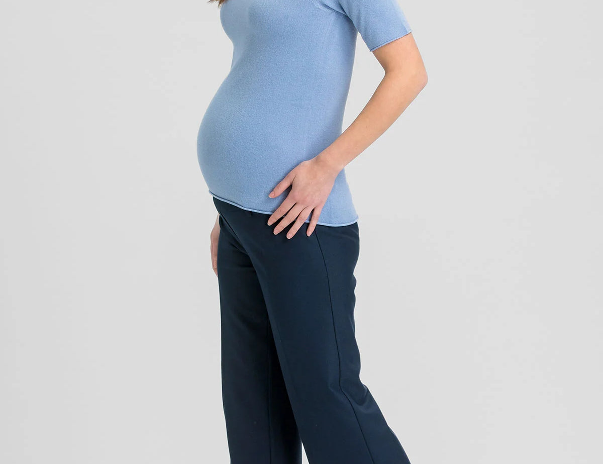 A pregnant woman posing in a light blue short-sleeve mock neck top and dark navy wide-leg trousers, paired with brown pointed-toe heels, against a solid light gray background.