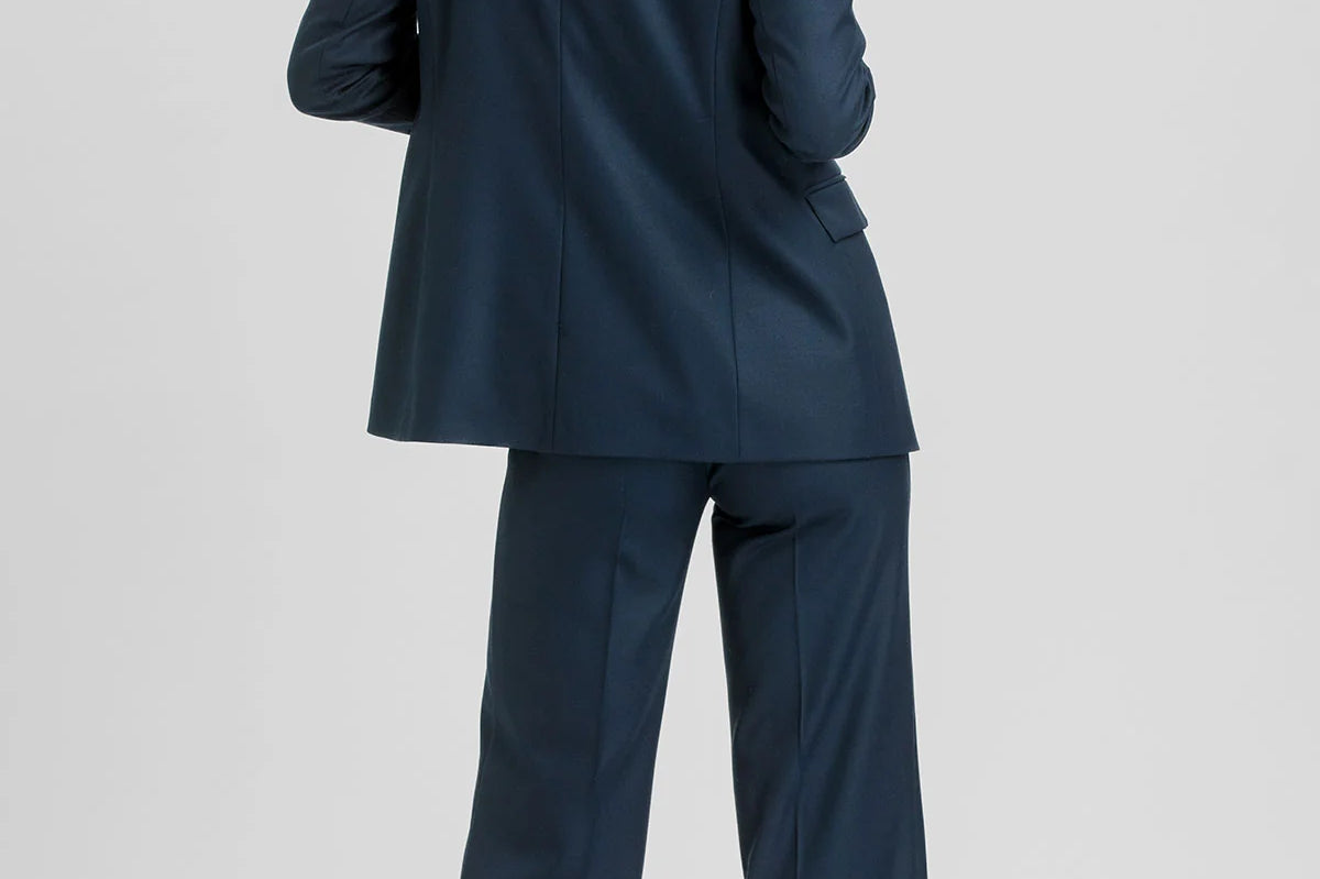 A woman wearing a navy blue two-piece suit, shown from the back, standing against a light gray studio background.