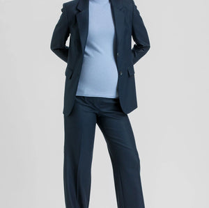 A woman modeling a navy blue two-piece suit consisting of a blazer and wide-leg trousers, worn over a light blue top and brown pointed-toe heels, against a light gray studio background.