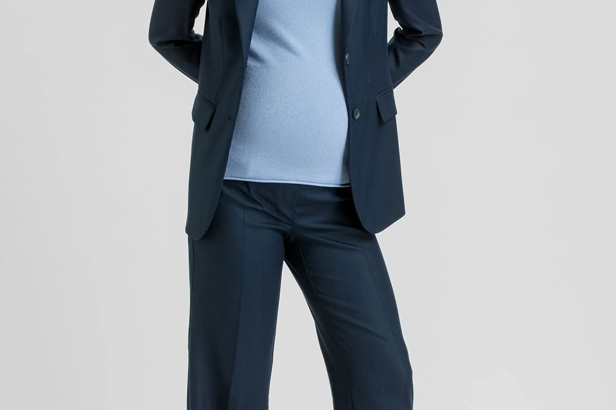 A woman modeling a navy blue two-piece suit consisting of a blazer and wide-leg trousers, worn over a light blue top and brown pointed-toe heels, against a light gray studio background.