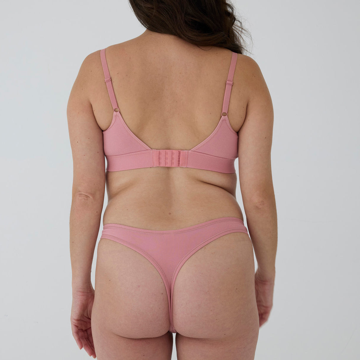 A woman shown from the back wearing a dusty pink ribbed bralette and matching thong against a plain light gray studio background.