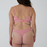 A woman shown from the back wearing a dusty pink ribbed bralette and matching thong against a plain light gray studio background.