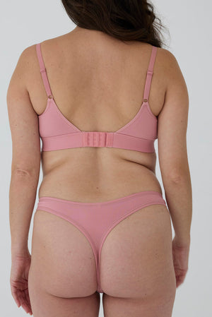 A woman shown from the back wearing a dusty pink ribbed bralette and matching thong against a plain light gray studio background.