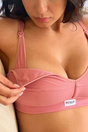 A close-up of a woman wearing a ribbed dusty pink wireless bra with a small white RENUF label on the band, sitting on a beige cushion.