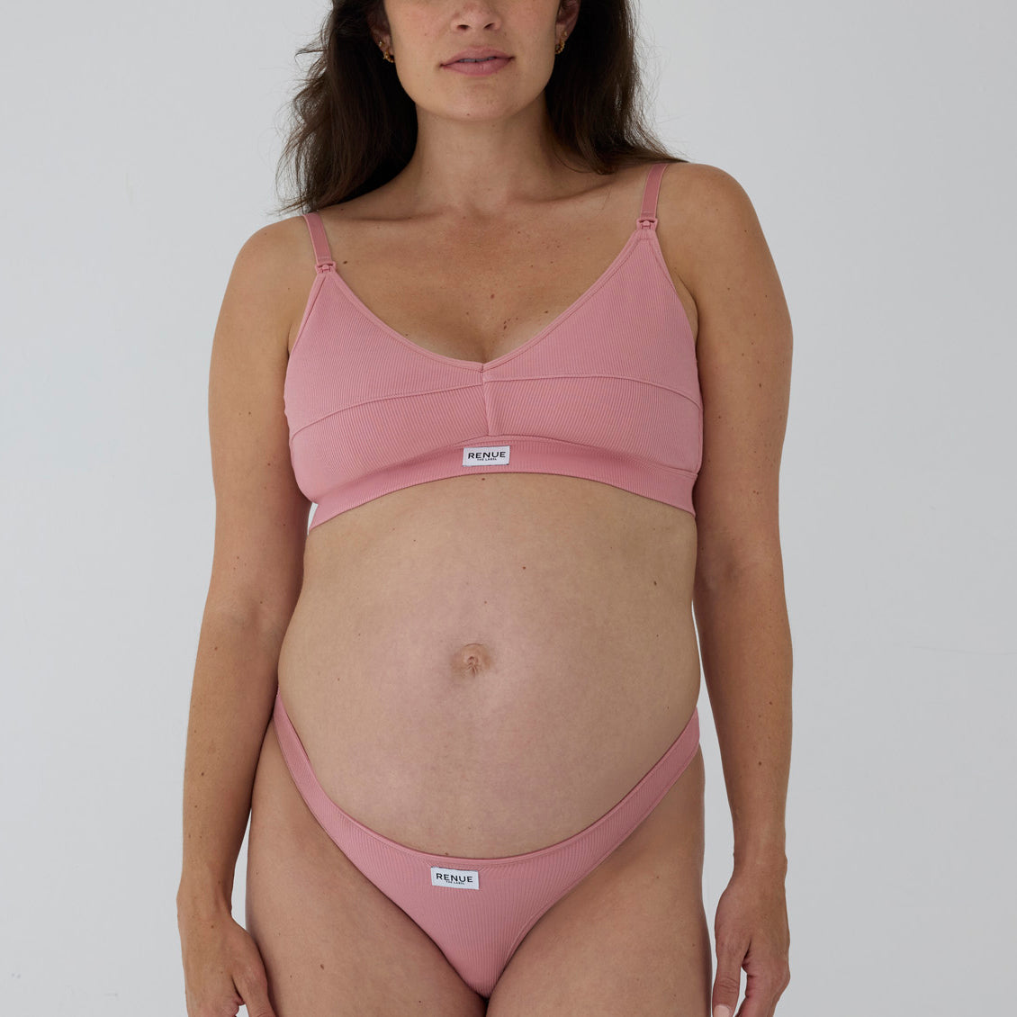 A pregnant model wearing a dusty pink maternity bralette and matching high-cut bikini bottoms by Nodusa, shown against a plain light gray studio background.