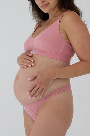 Pregnant model wearing a dusty pink maternity bra and matching underwear bottoms against a light gray studio background.