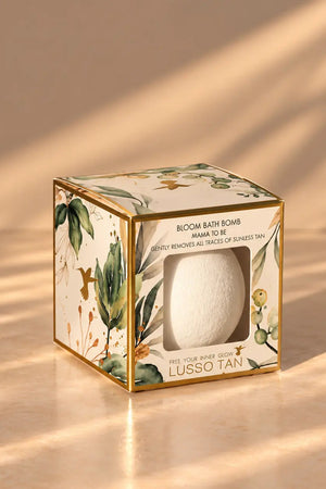 A boxed bath bomb product sits on a warm beige surface with soft diagonal shadows. The square package has botanical illustrations, gold trim, and a clear window showing a white bath bomb inside.