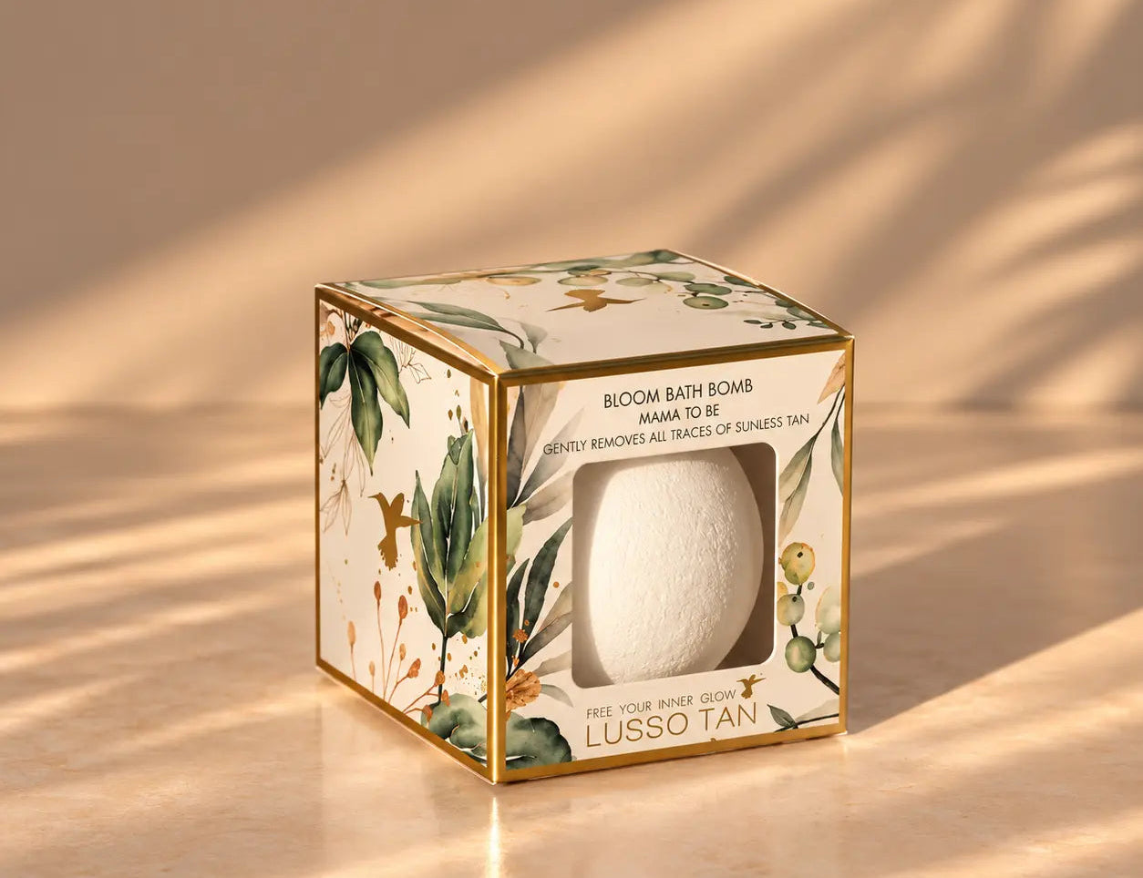 A boxed bath bomb product sits on a warm beige surface with soft diagonal shadows. The square package has botanical illustrations, gold trim, and a clear window showing a white bath bomb inside.