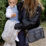 Woman holding a baby and carrying a gray handbag outdoors.